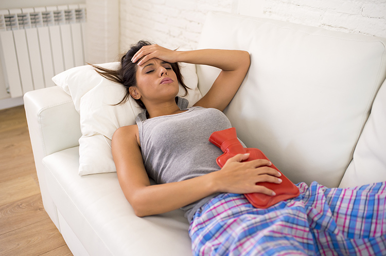 menstrual-cramping a woman lying down with a hot water bottle on her stomach and her hand placed on her forward
